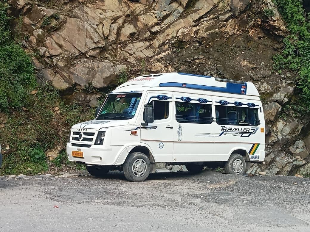 White Force Traveller high-roof van with a blue roof stripe and blue window trim, Traveller lettering on the side, a yellow commercial number plate and MISHRA lettered on the rear corner, parked on a gravel mountain road in front of a grey rock cutting with green slopes on either side